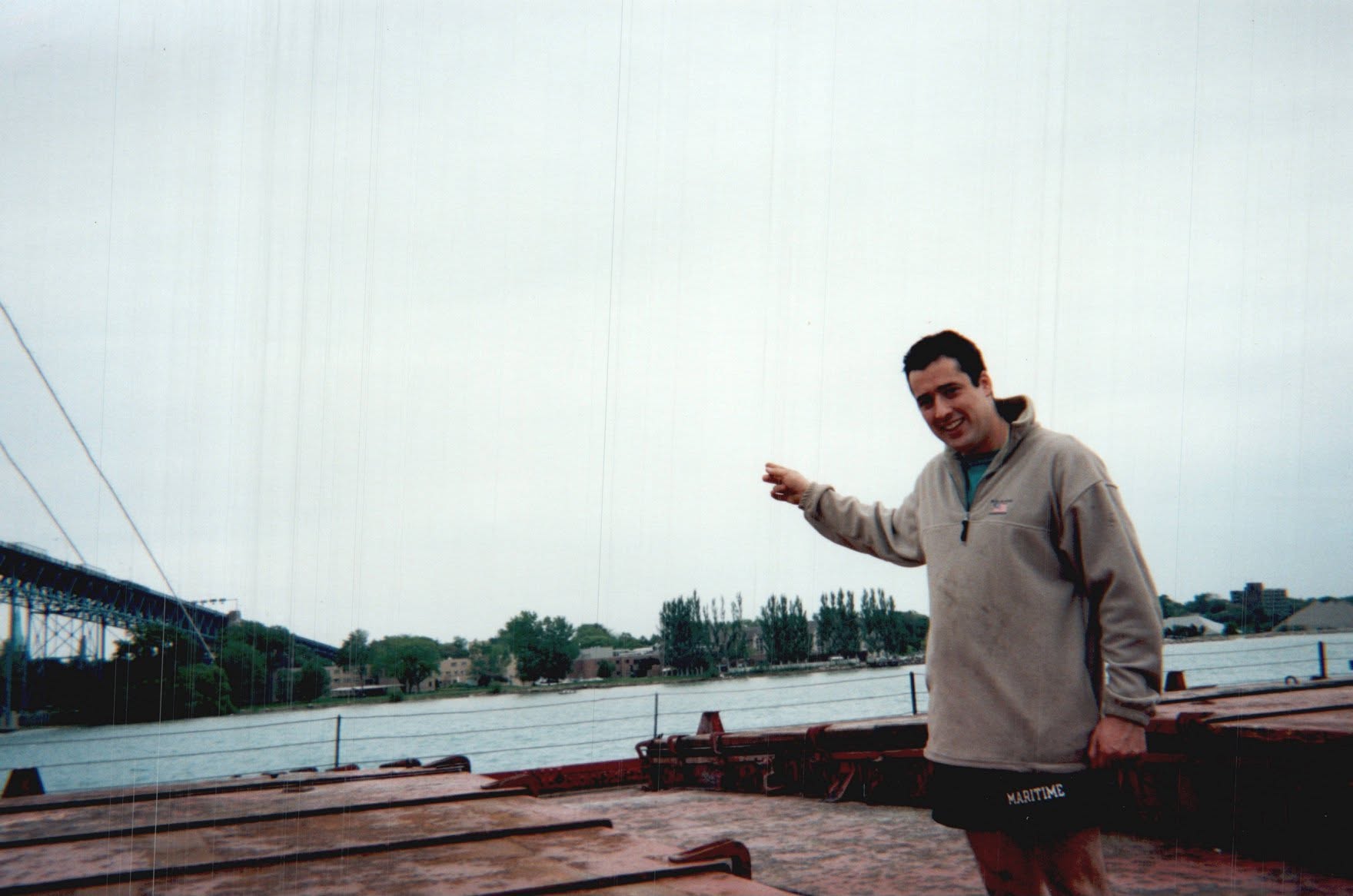 A man stands on the deck of a ship beside a river.