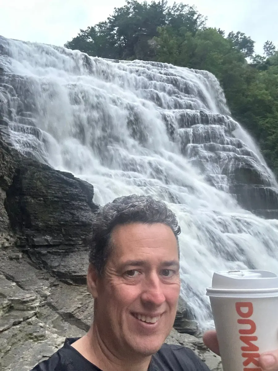 A man smiles beside a waterfall while holding a cup.